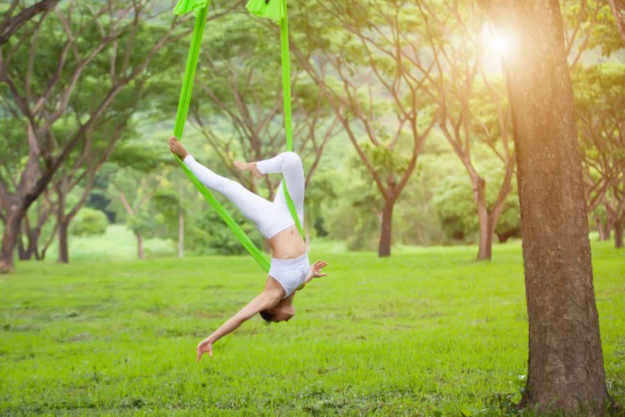 Person practising aerial yoga upside down in a green hammock outdoors, surrounded by trees and bright natural sunlight.