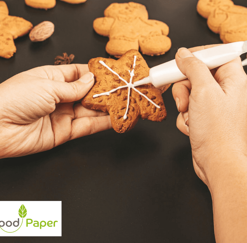 Hands piping icing on a star-shaped gingerbread biscuit, with Food Paper logo watermark, showcasing baking and decoration process.