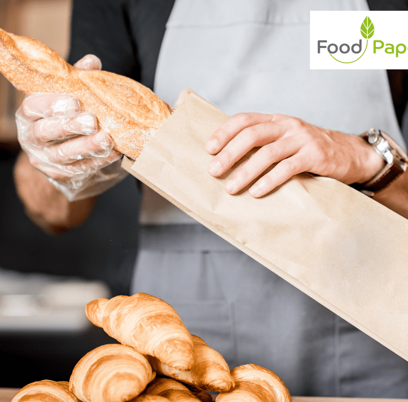 Baker placing fresh baguette into paper bag with croissants on counter, Food Paper logo watermark shown for branding.