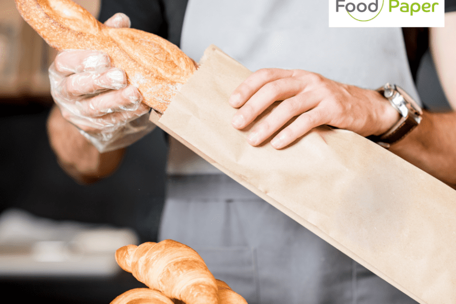 Baker placing fresh baguette into paper bag with croissants on counter, Food Paper logo watermark shown for branding.