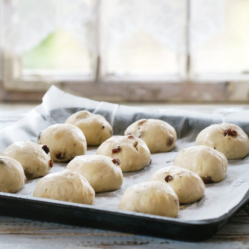 Fresh dough balls with raisins resting on a tray lined with non-stick silicone baking paper in a rustic kitchen setting.