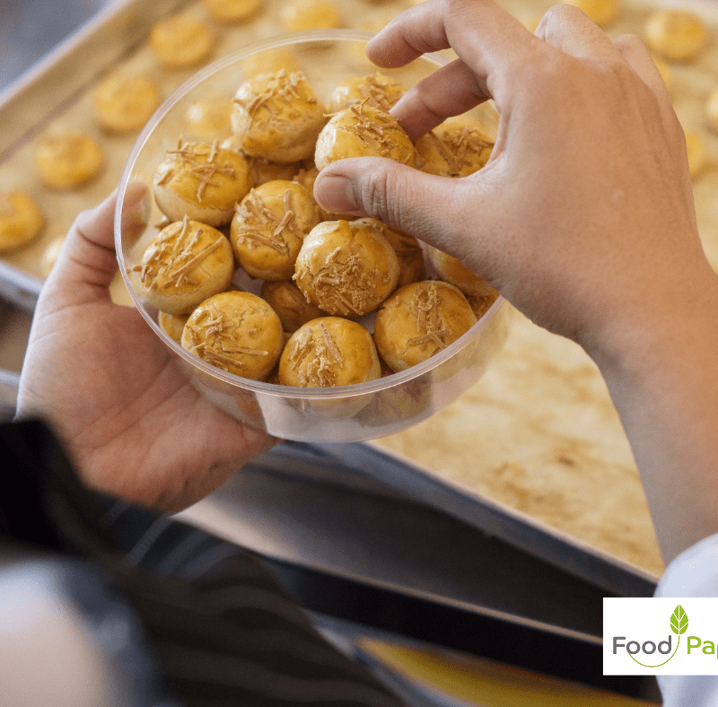 Hands placing baked cheese pastries into a clear container, Food Paper logo watermark, highlighting baking and packaging solutions.