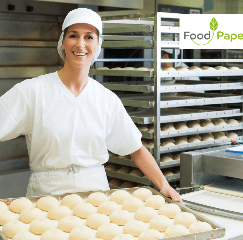 Female baker in white uniform holding tray of fresh bread rolls in bakery, Food Paper logo watermark in corner for branding.