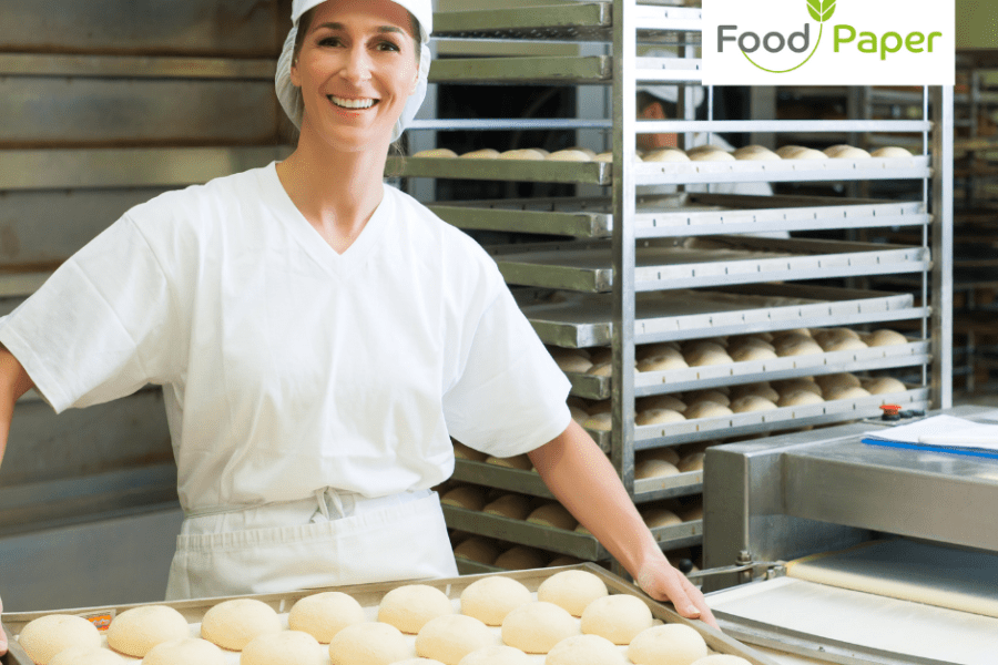 Female baker in white uniform holding tray of fresh bread rolls in bakery, Food Paper logo watermark in corner for branding.
