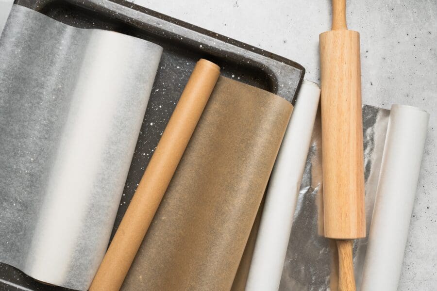 Rolls of baking paper, parchment, and foil with a wooden rolling pin and tray, essential for cooking and baking preparation.