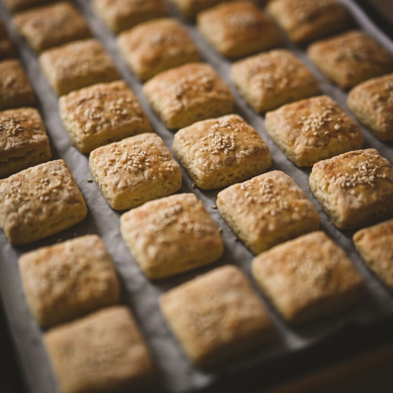 Golden square biscuits topped with sesame seeds, freshly baked on parchment paper in a UK bakery tray.