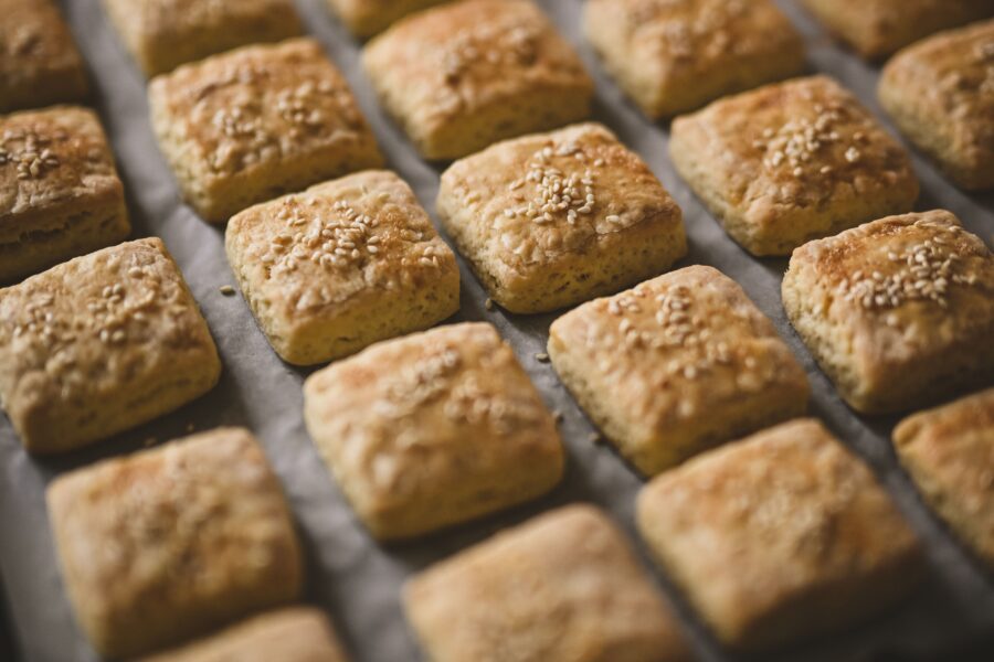 Golden square biscuits topped with sesame seeds, freshly baked on parchment paper in a UK bakery tray.