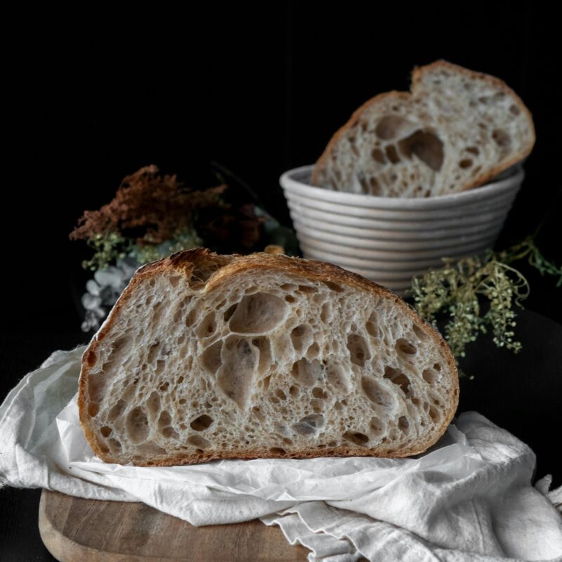 Half sourdough loaf on a wooden board with cloth, showing airy open crumb and crisp crust, styled with rustic bakery elements.