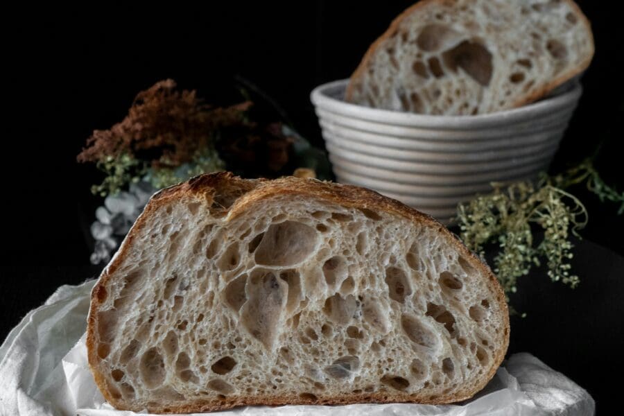 Half sourdough loaf on a wooden board with cloth, showing airy open crumb and crisp crust, styled with rustic bakery elements.